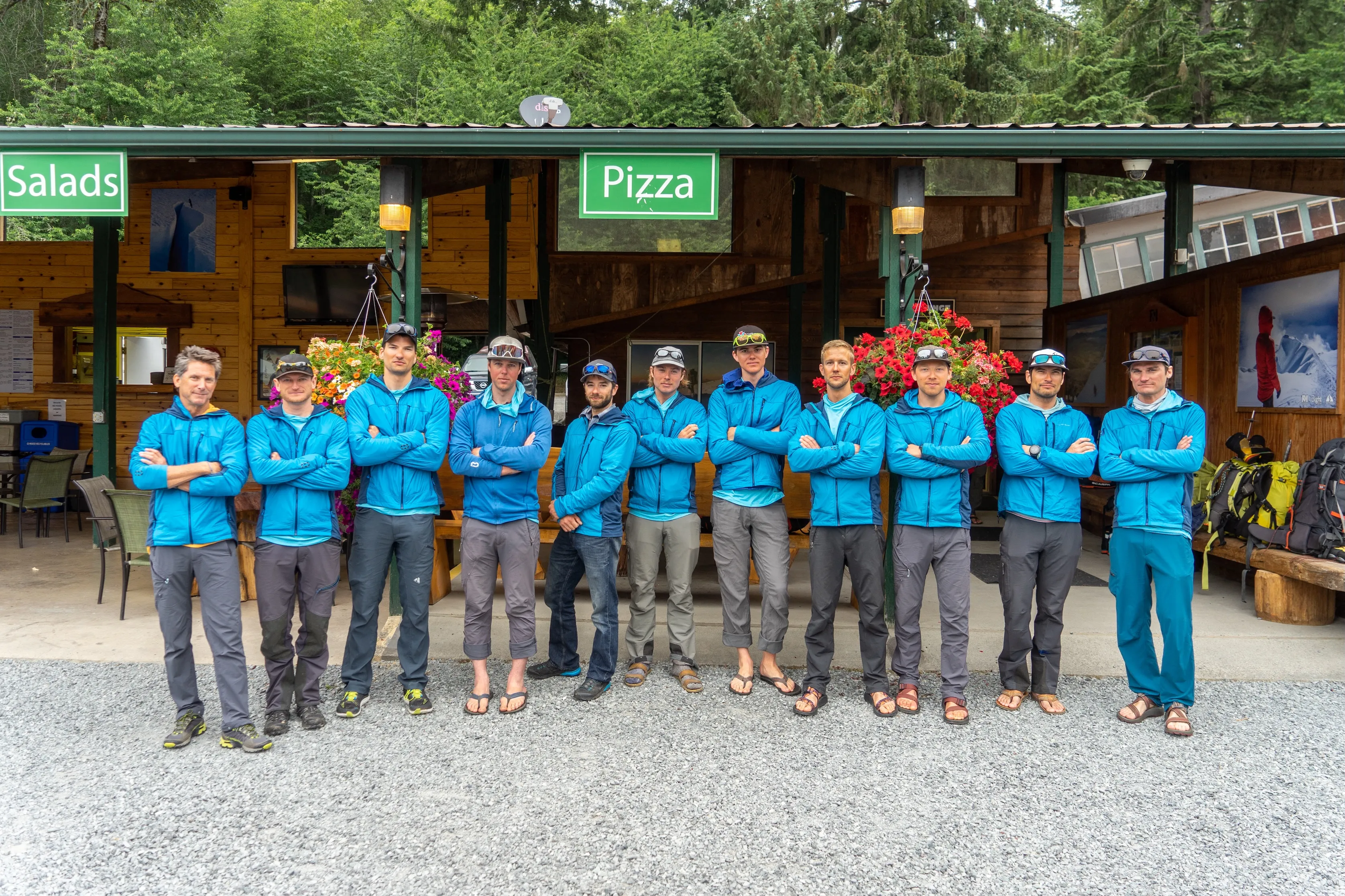 Eleven men in blue jackets stand with arms crossed in front of a wooden building with signs for salads and pizza.