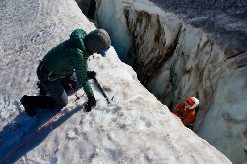 Two climbers navigating snowy terrain next to a deep icy crevasse, one assisting the other.