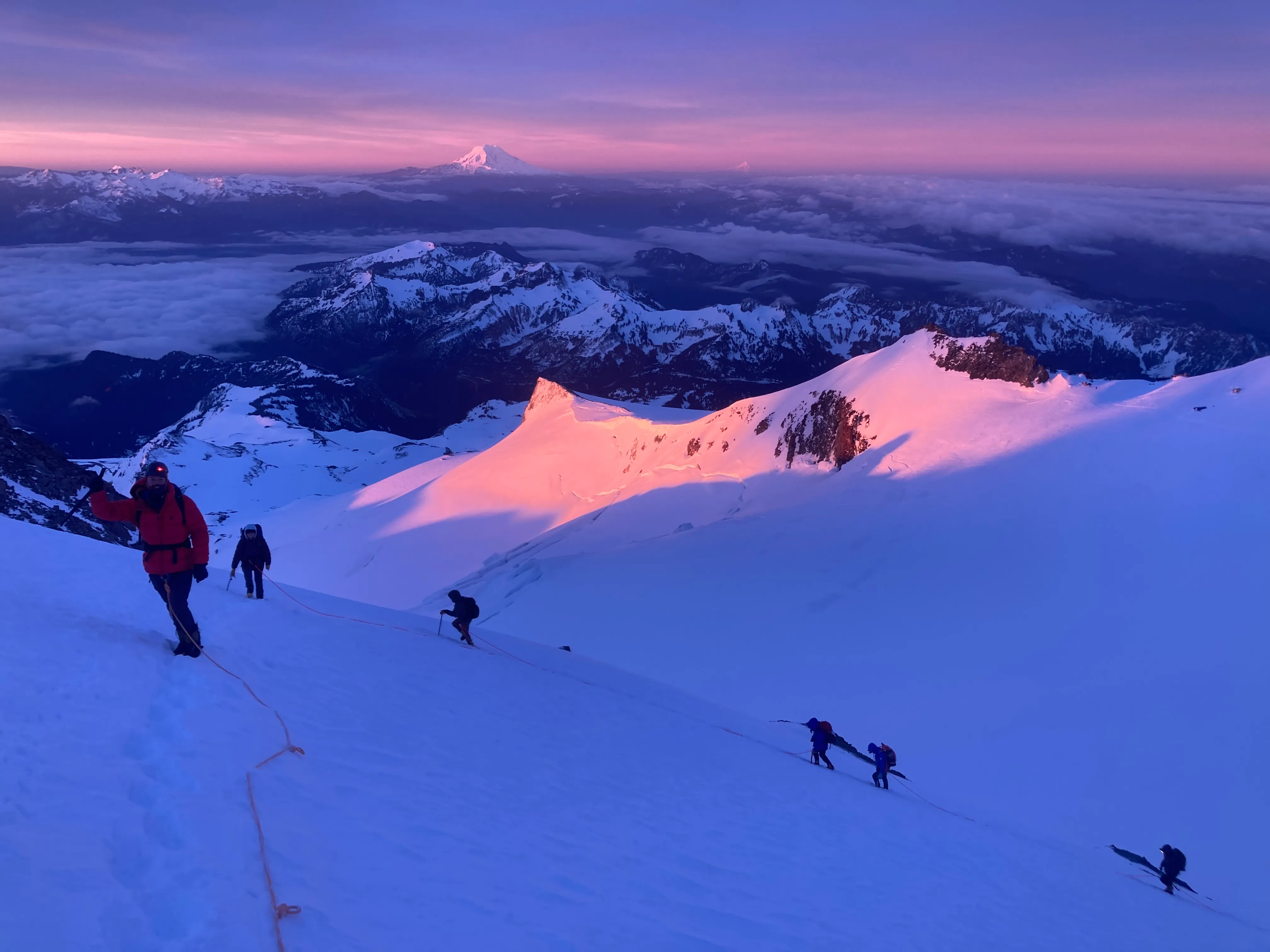 Climbers roped together ascending snowy mountain slope at sunrise with distant peaks and purple sky.