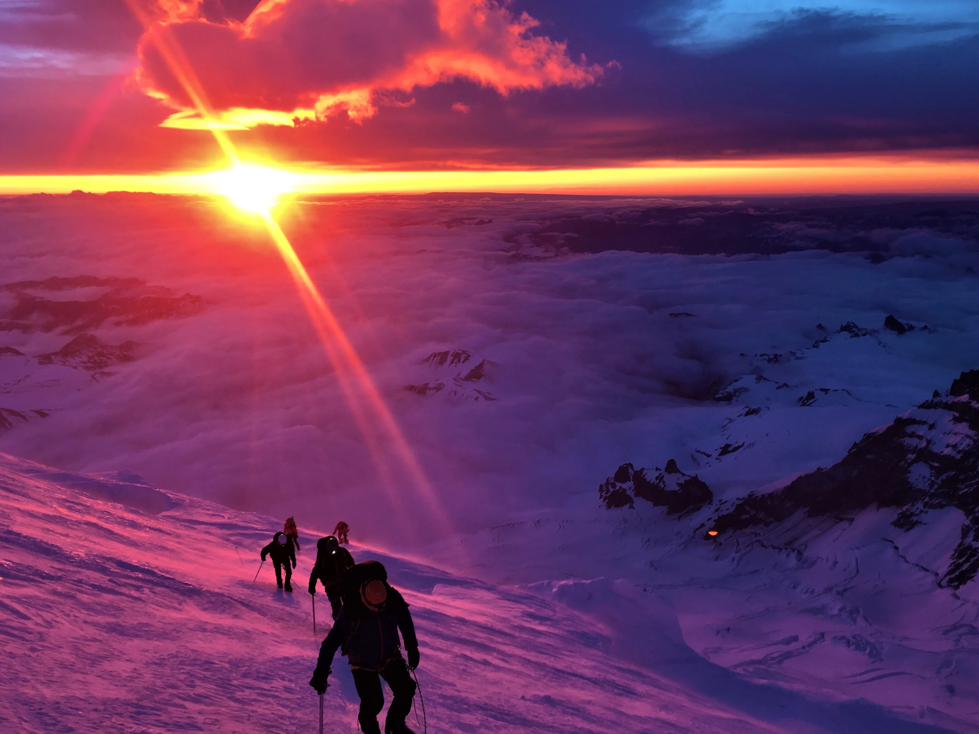 Climbers ascending a snowy mountain slope at sunrise with clouds and mountains below.