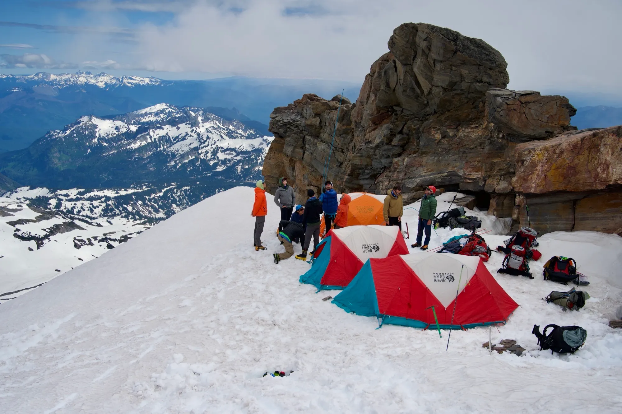 Group camping on snowy mountain slope with colorful tents and rocky backdrop under cloudy sky.