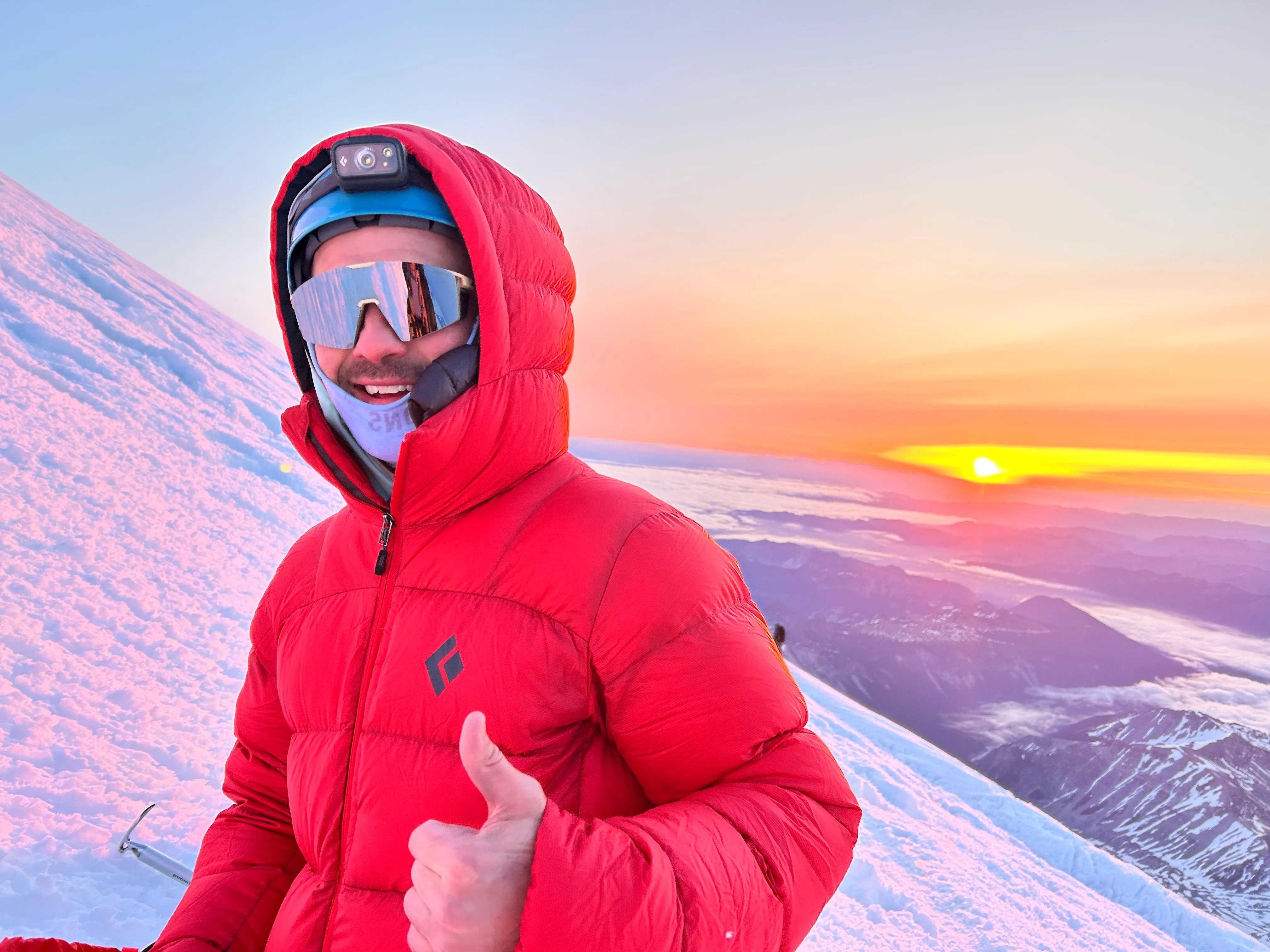 Person in red jacket and helmet with headlamp giving thumbs up on snowy mountain at sunrise.