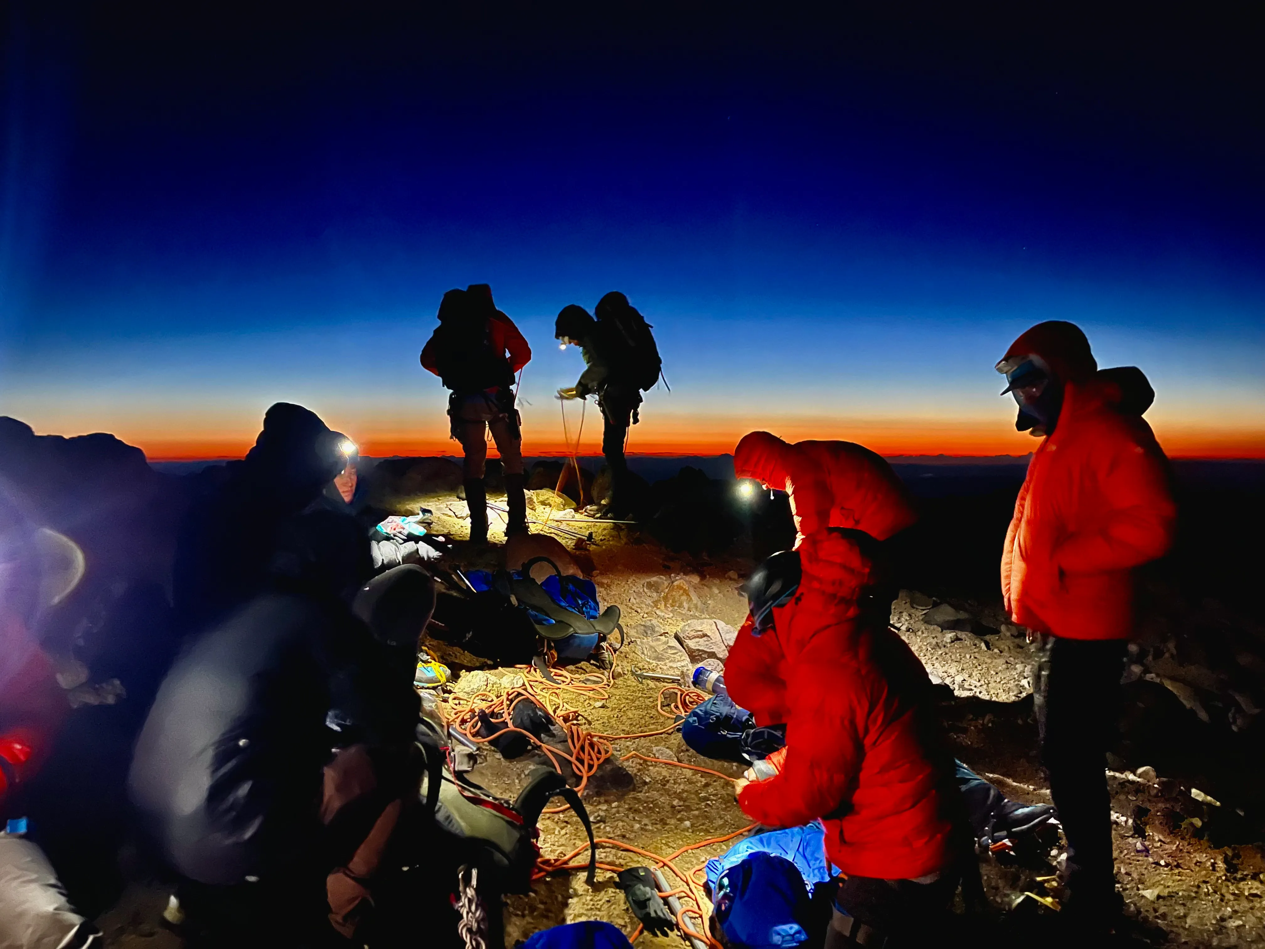 Climbers wearing headlamps and red jackets preparing gear at dawn on rocky terrain.