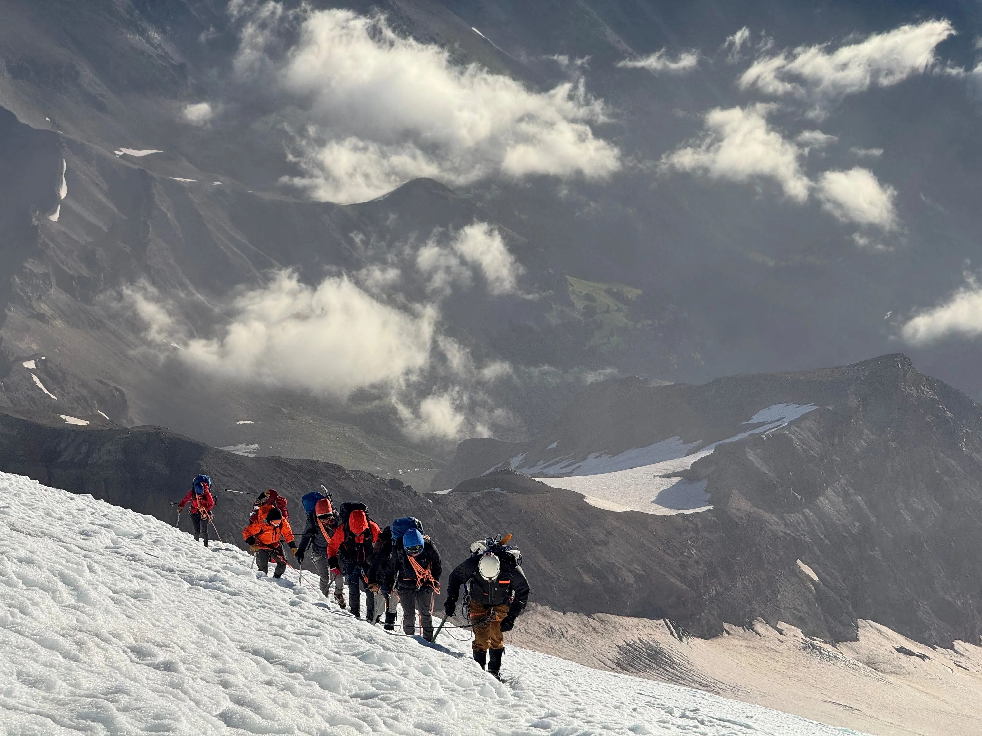 A group of climbers roped together ascending a snowy mountain slope under cloudy skies.