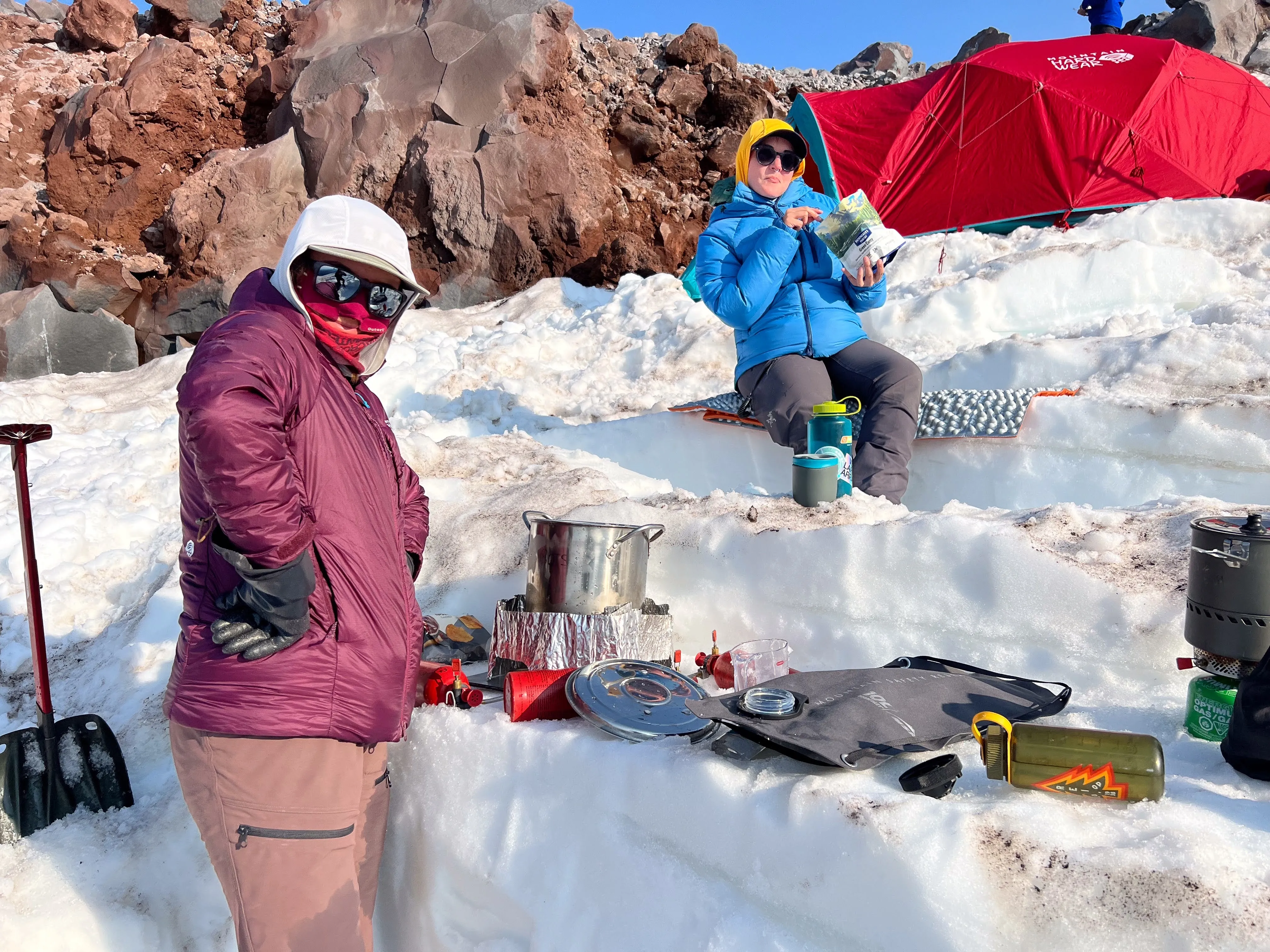 Two people in winter gear at a snowy campsite with cooking equipment and a red tent nearby.