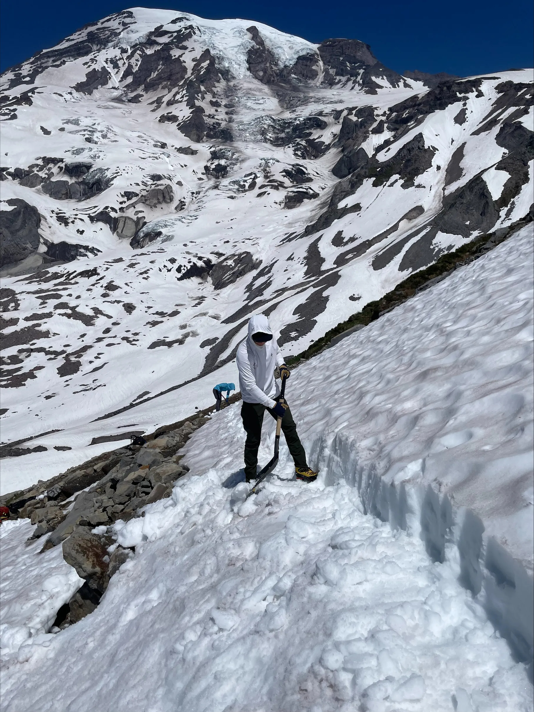 Person in white hoodie using a shovel on a snowy mountain slope with rocky patches and clear blue sky.