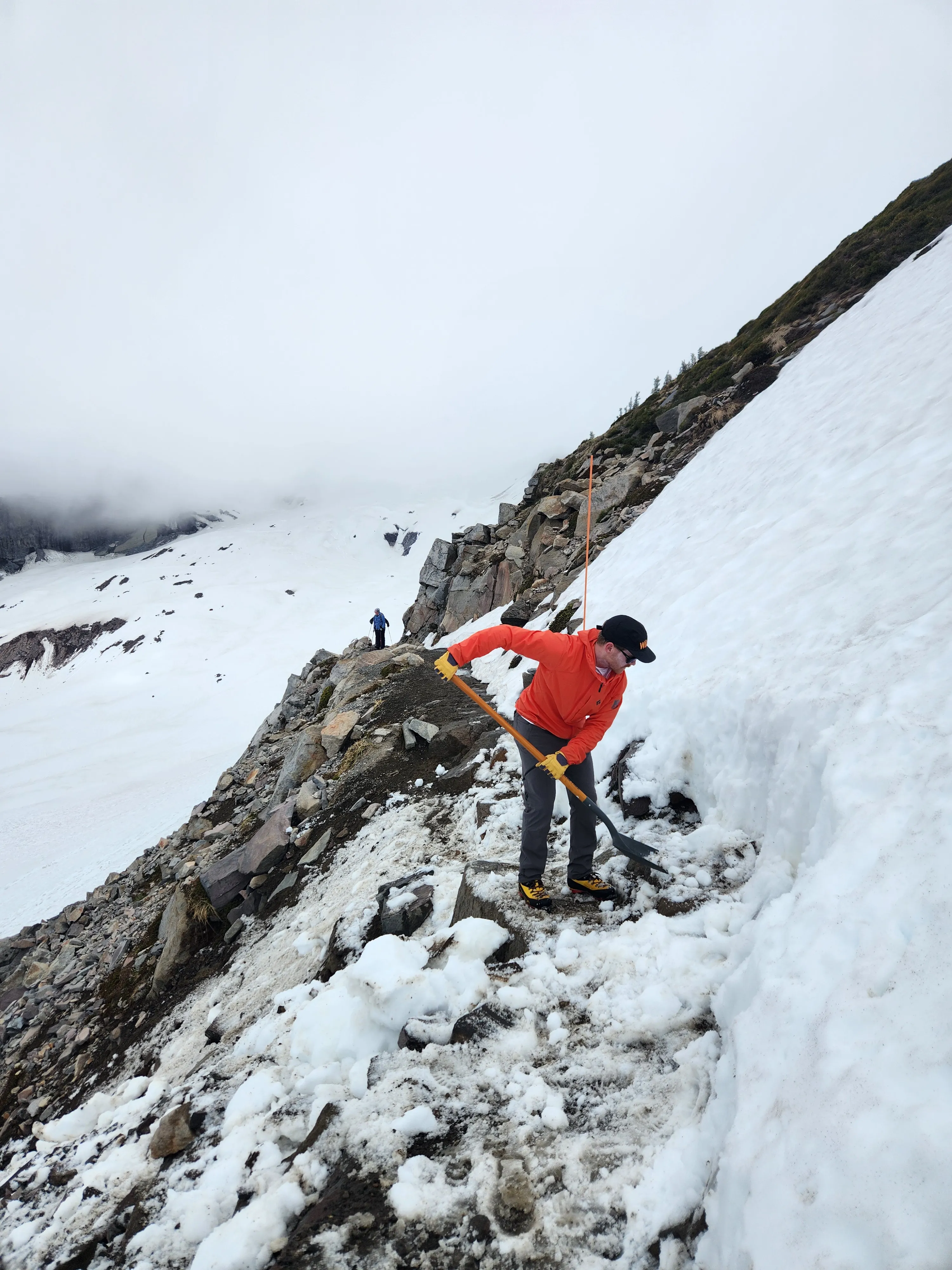 Person in orange jacket shoveling snow on a rocky mountain slope with another hiker in the background.
