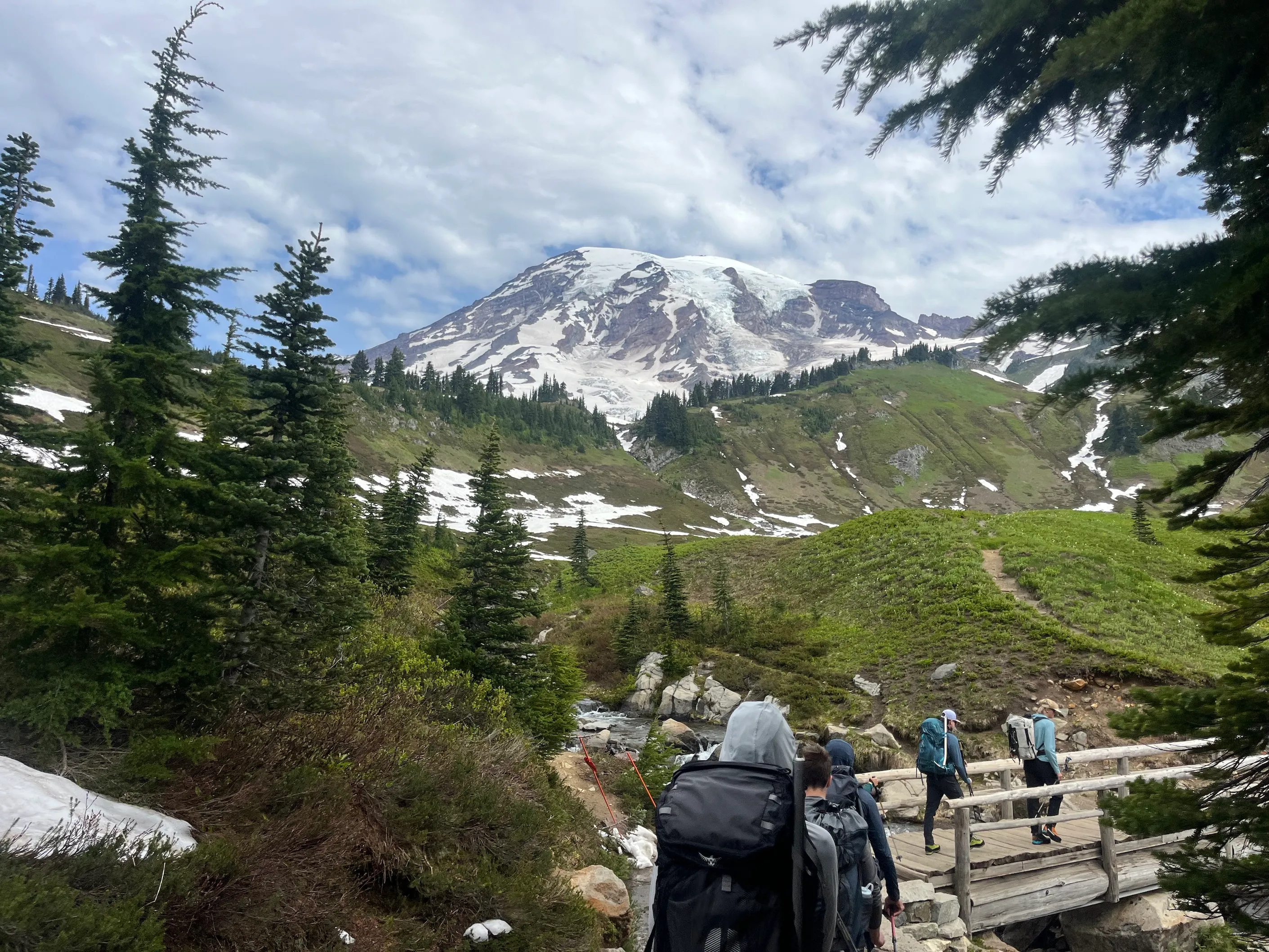 Hikers with backpacks crossing a wooden bridge with snow patches and a snow-capped mountain in background.