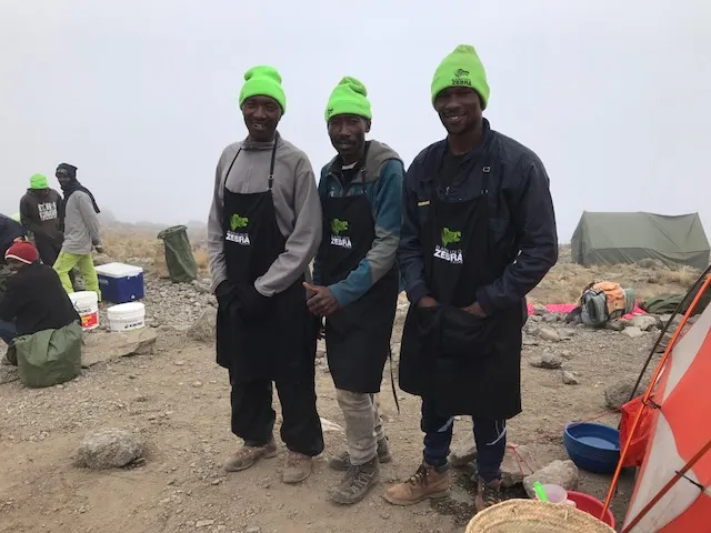 Three men in neon green hats and black aprons standing on rocky ground with tents in the foggy background.