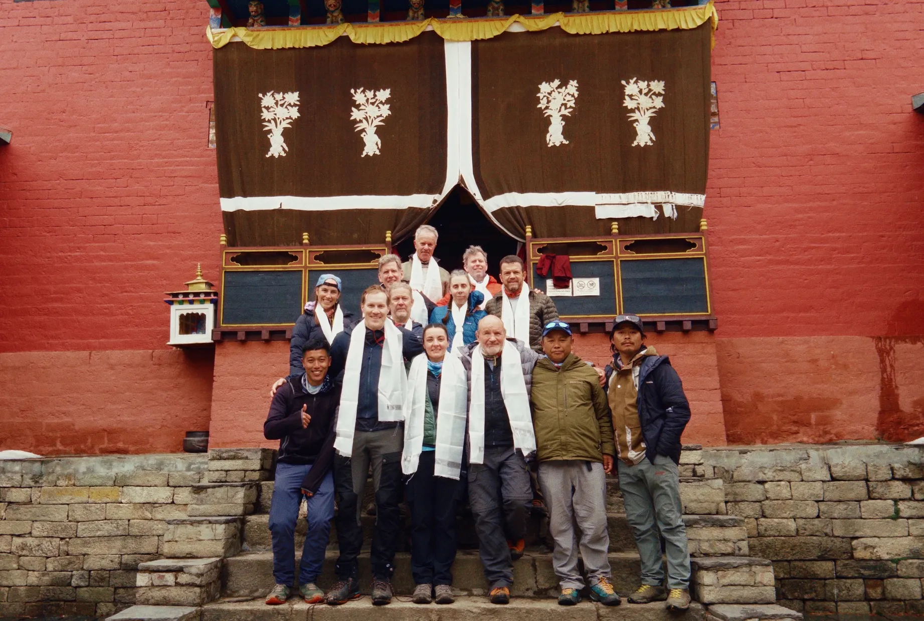 Group of hikers wearing white scarves posing on stone steps in front of red temple with decorated brown curtains.