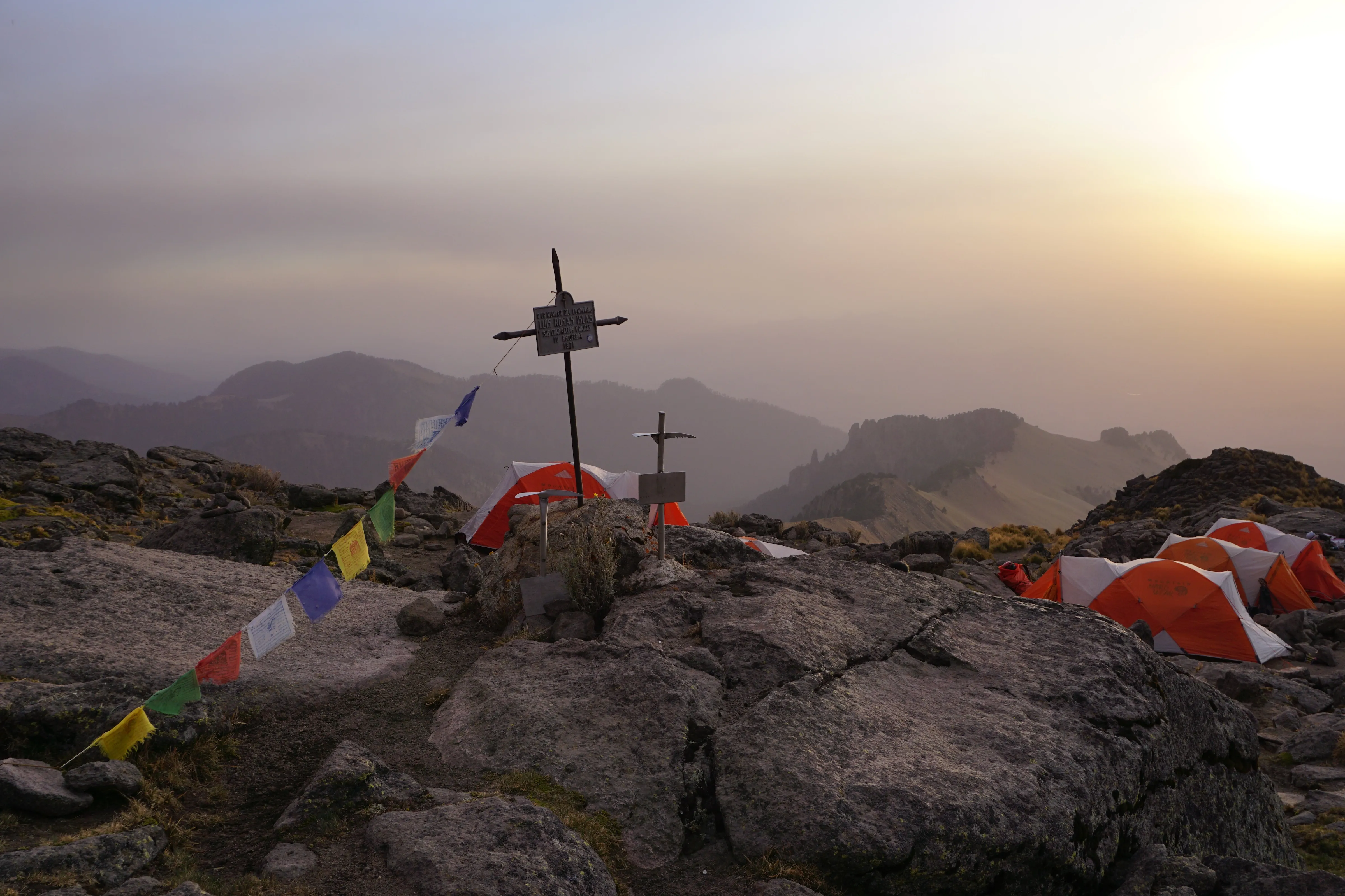 Mountain campsite with orange tents near a memorial cross at sunset with hazy hills in the background.