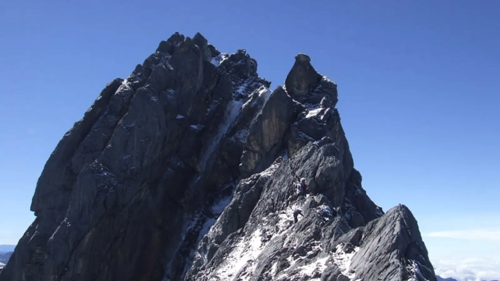 Climbers ascending a steep rocky mountain ridge with patches of snow under a clear blue sky.