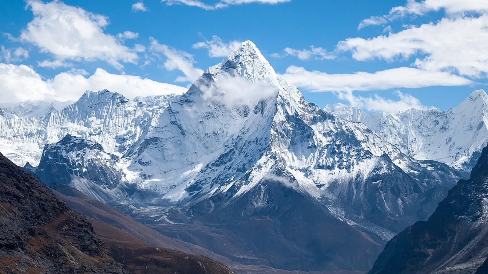 Snow-covered mountain peak with rocky slopes under blue sky and scattered clouds.