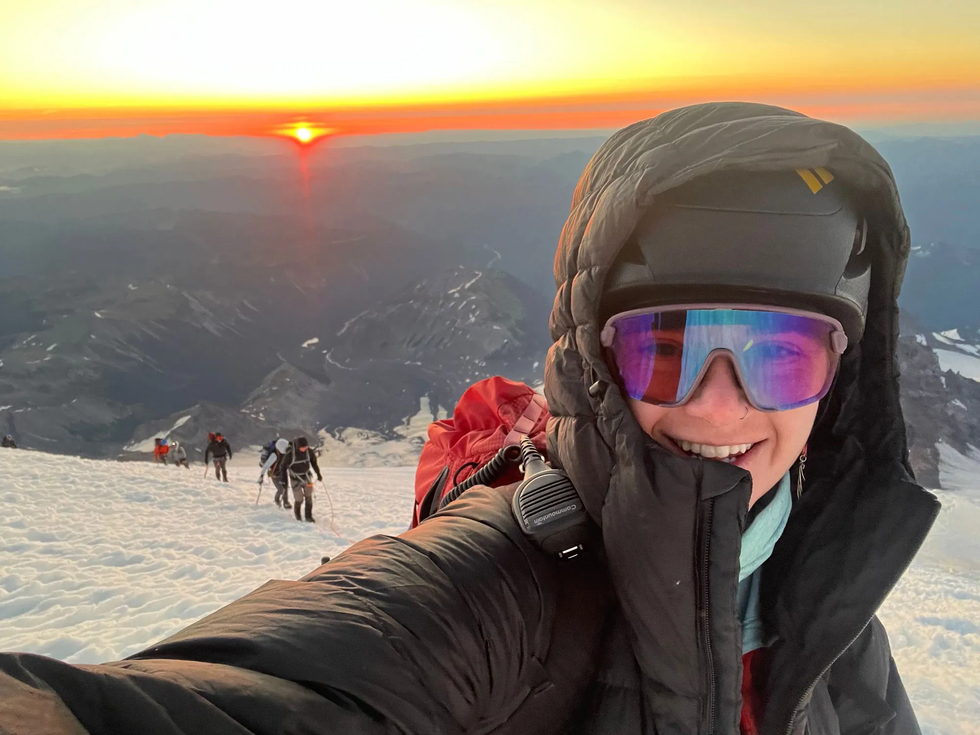 Smiling climber in winter gear takes selfie on snowy mountain at sunrise with others climbing behind.