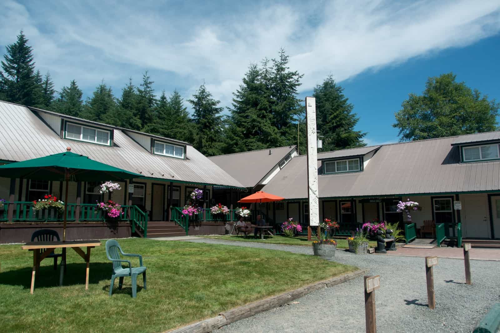 Green building with porch, flower pots, umbrellas, chairs, and tall trees under a blue sky.