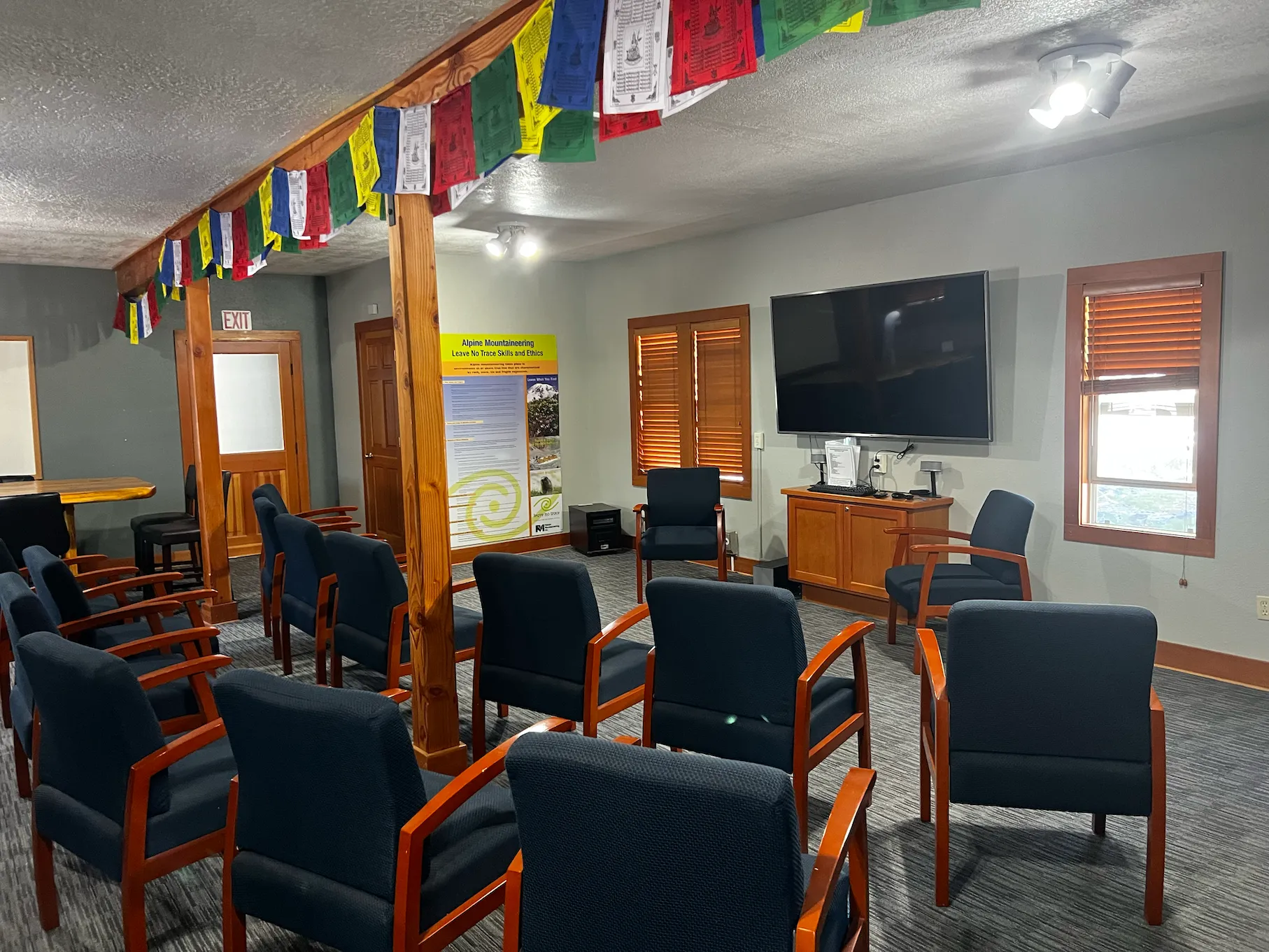Room with arranged dark blue chairs, wooden beams, prayer flags, a large TV, and a poster on the wall.