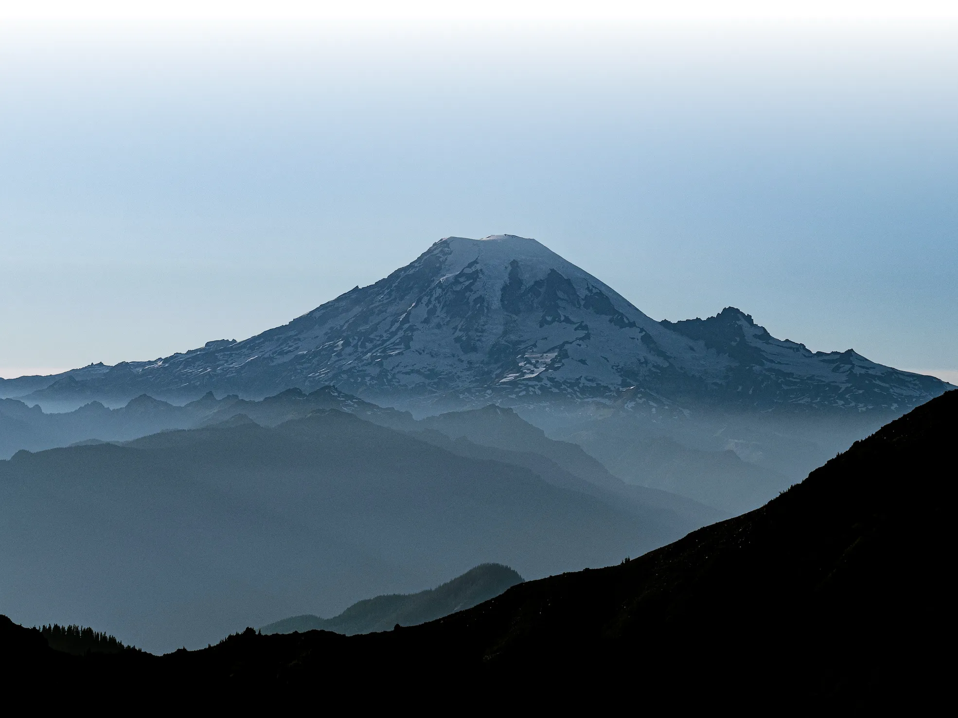 Snow-capped mountain with layered dark hills in the foreground under a clear sky.